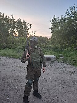 Airsoft player wearing a loaded plate carrier and chest rig near a garage